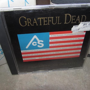 May include: A Grateful Dead CD case with a black cover. The text "Grateful Dead" is in gold. The case features a blue and red flag design with the letters "AcS" and the text "New Mexican Dreams". A sticker is attached.