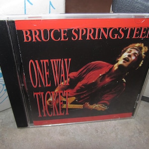 May include: A compact disc case featuring Bruce Springsteen's album "One Way Ticket." The album cover has a black background with red text and a photo of Springsteen playing guitar and singing.