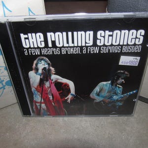 May include: A music CD in a clear plastic jewel case featuring The Rolling Stones. The cover art displays a live concert photograph with the text "THE ROLLING STONES" and "A FEW HEARTS BROKEN, A FEW STRINGS BUSTED" printed in white against a black background.