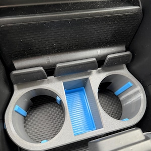 Op de afbeelding: Een grijze plastic bekerhouder-inzet voor in de auto met twee ronde openingen met blauwe rubberen grepen en een centraal rechthoekig opbergvak met een geribbelde blauwe bodem, onder een consolepaneel met twee USB-poorten en een AUX-ingang.