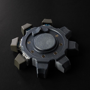 May include: A grey and blue gear-shaped object with a central circular design and eight protruding sections. The object has a textured surface and small metallic accents. The background is solid black.