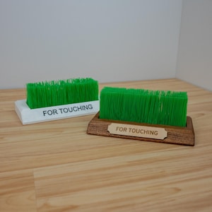 May include: Two rectangular objects with green bristles, resembling miniature lawns. One is in a white base, the other in a wooden base. Both have the text "FOR TOUCHING" printed on them. The objects are on a light wood surface.