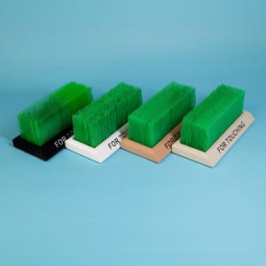 Op de afbeelding: Vier rechthoekige borstels met groene borstelharen en schuine bases. De bases zijn zwart, wit, bruin en wit, elk met de tekst "FOR TOUCHING". De borstels zijn gerangschikt op een lichtblauwe achtergrond.