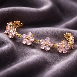 May include: A gold-toned bracelet featuring pink flower-shaped gemstones. The bracelet is set against a dark gray, velvety background, highlighting the delicate design and sparkle of the jewelry.