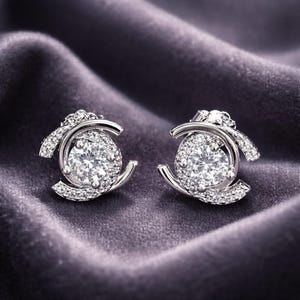 May include: A pair of silver-toned stud earrings. Each earring features a large, round, clear gemstone surrounded by smaller stones and a curved, open design. The earrings are set against a dark purple, textured background.