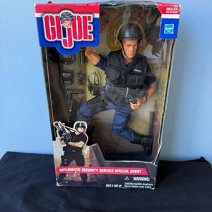 May include: A G.I. Joe Diplomatic Security Service Special Agent action figure in original packaging. The figure wears a black tactical vest, blue pants, and a helmet, holding a black submachine gun. Accessories include a pistol and headset.