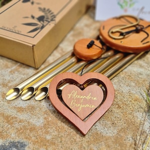 May include: A wind chime featuring gold-toned metal tubes, a round wooden top, and a wooden heart-shaped pendant. The pendant has a smaller heart cutout with the gold-coloured text Alexandria & Benjamin printed on it. A cardboard box is visible nearby.