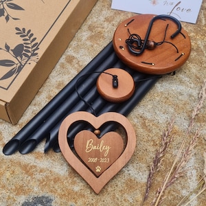May include: A wooden wind chime with a heart-shaped pendant engraved with the name "Bailey" and dates 2008-2023, along with a paw print. The chime features black tubes, a wooden top, and a wooden clapper. The chime is packaged in a cardboard box.