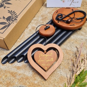 May include: A black metal wind chime with wooden heart-shaped ornaments. The heart has a smaller heart cutout with the inscription "Margaret Ann Smith 1935-2023" and a small cross.