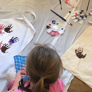 May include: A child is making handprints on a white tote bag and a white apron. The tote bag and apron have brown, purple, and pink handprints. There are paint tubes and a paint palette on a white plastic tablecloth.