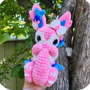 Könnte beinhalten: Handgemachtes rosa Sylvy das kuschelige Schwanzmonster-Plüschtier mit blauen und weißen Akzenten. Das Plüschtier hat große Ohren, eine rüsselartige Nase und einen flauschigen Schwanz. Der Text "Sylvy das kuschelige Schwanzmonster" steht oben im Bild.