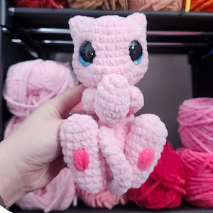 Könnte beinhalten: Ein rosa gehäkeltes Mew-Plüschtier mit blauen Augen und rosa Akzenten. Das Bild enthält den Text "Meu the cuddly tail monster" und "Pattern by Saren Bowie s_c.handmade". Im Hintergrund sind verschiedene Wollfarben zu sehen.