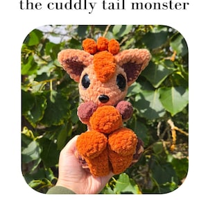 Op de afbeelding: Een gehaakt pluche monster speelgoed genaamd Vulx the cuddly tail monster. Het wezen is gemaakt van oranje en bruin garen, heeft grote zwarte ogen, een kleine zwarte neus en een pluizige staart. Tekst: Vulx the cuddly tail monster. Pattern by Saren Bowie s_c.handmade.