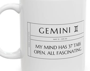 May include: A white ceramic mug featuring a black graphic design with the text Gemini, the dates May 21 - Jun 20, and the phrase My mind has 37 tabs open. All fascinating. The design includes a vertical label reading Zodiac Collection.