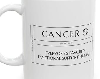 May include: A white ceramic mug featuring a black printed rectangular label. The text reads CANCER, the zodiac symbol, JUN 21 - JUL 22, and EVERYONE'S FAVORITE EMOTIONAL SUPPORT HUMAN. The side text reads ZODIAC COLLECTION.