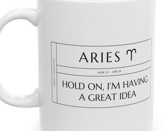 May include: A white ceramic mug featuring a black graphic design. The text reads ARIES with the ram symbol, MAR 21 - APR 19, and HOLD ON, I'M HAVING A GREAT IDEA. A vertical label on the left side of the box reads ZODIAC COLLECTION.