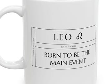 May include: A white ceramic mug featuring a black rectangular graphic with the text LEO, the Leo zodiac symbol, the dates JUL 23 - AUG 22, and the phrase BORN TO BE THE MAIN EVENT. The side of the graphic includes the text ZODIAC COLLECTION.