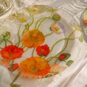 Può includere: Tre piatti di vetro trasparente con un motivo floreale di papaveri arancioni, rossi e gialli. I piatti sono disposti su una tovaglia bianca, con un bicchiere d'acqua e posate.