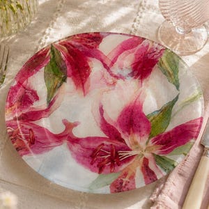Può includere: Un piatto decorativo con un motivo floreale con gigli rosa e bianchi e foglie verdi. Il piatto è appoggiato su una tovaglia bianca, accanto a un bicchiere, posate e una candela in un portacandele di vetro. Un bouquet di fiori è sullo sfondo.