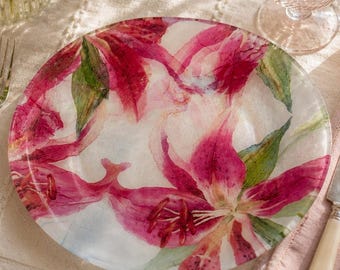 Könnte beinhalten: Ein dekorativer Teller mit einem Blumendesign mit rosa und weißen Lilien und grünen Blättern. Der Teller steht auf einer weißen Tischdecke, zusammen mit einem Glas, Besteck und einer Kerze in einem Glashalter. Ein Blumenstrauß ist im Hintergrund.