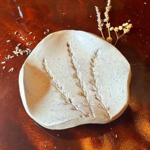 Puede incluir: Un plato decorativo de cerámica en color blanco roto con forma irregular y un diseño en relieve de ramas de lavanda. La superficie presenta sutiles motas púrpuras y el plato se acompaña de una pequeña rama de lavanda seca.