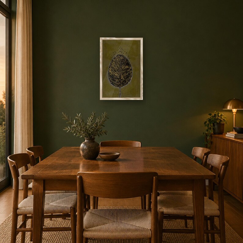 May include: A framed wall art print featuring a dark, textured leaf illustration with gold accents on an olive green background, displayed in a light-colored frame centered on a dark green wall above a wooden dining table.