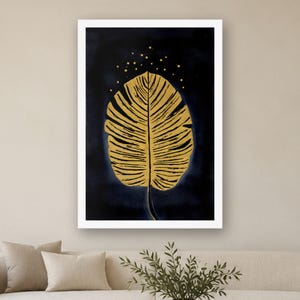 May include: A framed wall art print featuring a large, textured golden leaf centered against a deep navy blue background. Above the leaf, small golden dots are scattered, resembling stars. The artwork is enclosed in a clean white frame.