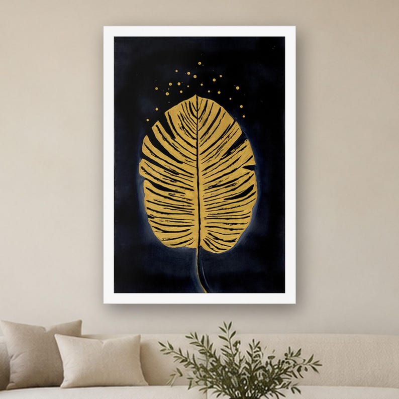 May include: A framed wall art print featuring a large, textured golden leaf centered against a deep navy blue background. Above the leaf, small golden dots are scattered, resembling stars. The artwork is enclosed in a clean white frame.