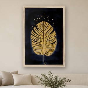 May include: A framed wall art print featuring a large, golden, textured leaf centered against a deep navy blue background. Above the leaf, small golden dots are scattered, creating a subtle speckled effect within the dark space.