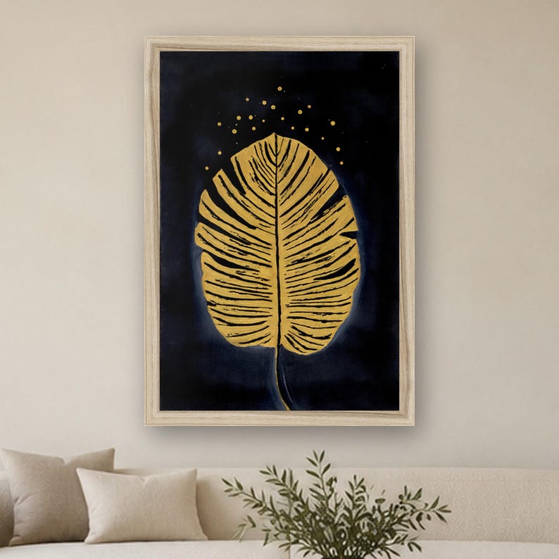 May include: A framed wall art print featuring a large, golden, textured leaf centered against a deep navy blue background. Above the leaf, small golden dots are scattered, creating a subtle speckled effect within the dark space.