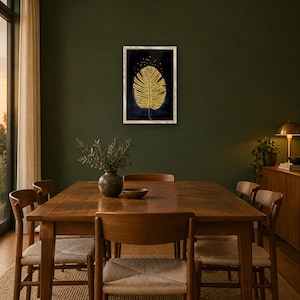 May include: A rectangular wall art piece in a light-colored frame featuring a gold-colored monstera leaf illustration centered on a dark navy blue background with small gold dots above the leaf, displayed on a dark green wall.