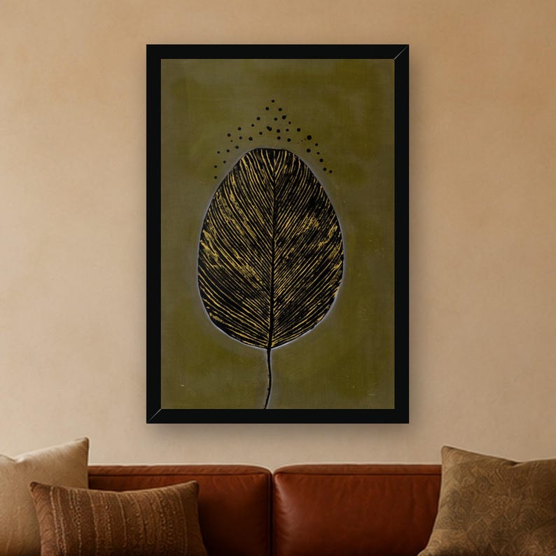 May include: A framed wall art print featuring a large, stylized black leaf with gold-toned vein details centered on a muted olive green background. Above the leaf, a small arc of black dots is arranged in a symmetrical pattern.