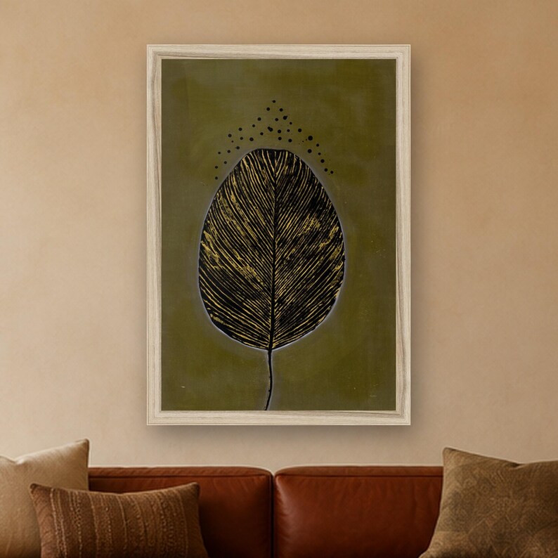 May include: A framed wall art print featuring a large, detailed black and gold leaf illustration centered on a muted olive green background. Above the leaf, a small arc of black dots is arranged in a symmetrical pattern. The artwork is enclosed in a light-colored wooden frame.