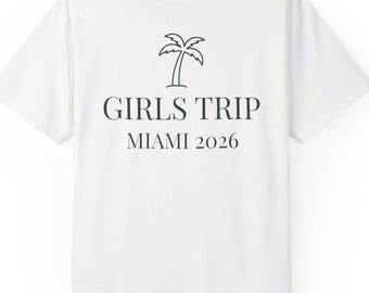 May include: A white short-sleeved t-shirt featuring a black line-art palm tree icon centred above the text GIRLS TRIP and MIAMI 2026 in a dark serif font. The shirt has a crew neckline and a visible brand label at the inner collar area.