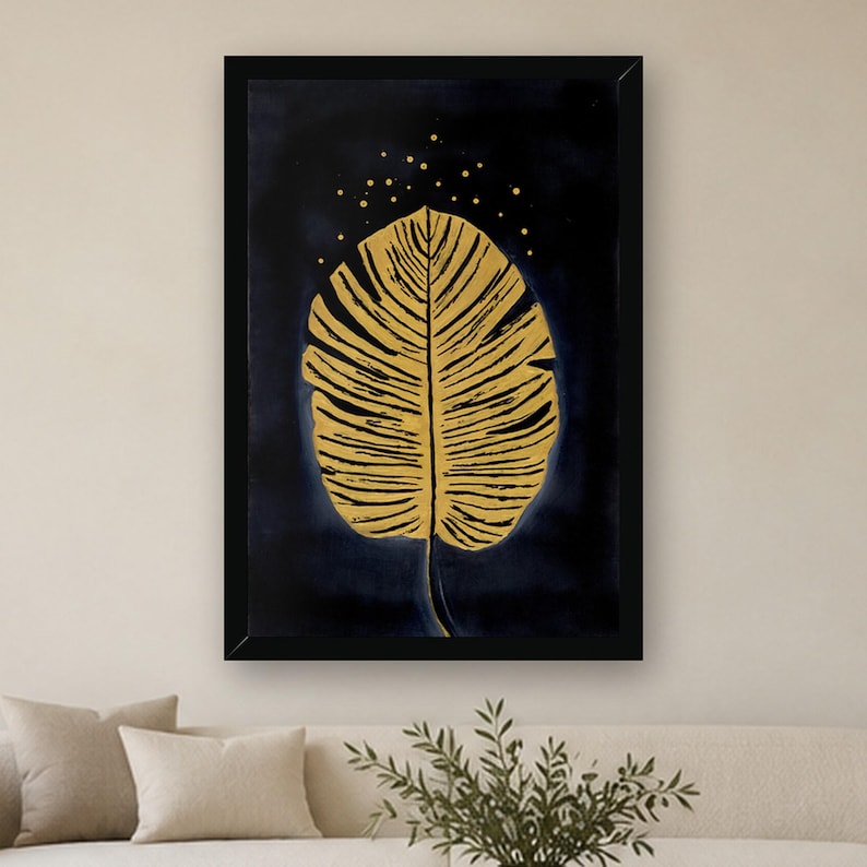 May include: A framed wall art print featuring a large, detailed golden leaf centered against a deep navy blue background. Above the leaf, small golden dots are scattered, creating a subtle speckled effect within the black frame.
