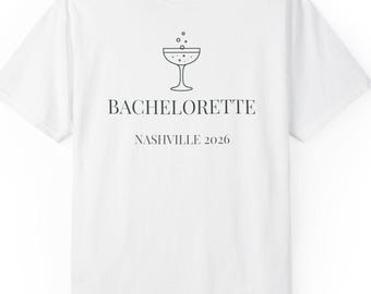 May include: A white short-sleeved t-shirt featuring a black line drawing of a stemmed cocktail glass with bubbles above it. Below the graphic, the text reads BACHELORETTE in a bold serif font, followed by the text NASHVILLE 2026 in a smaller font.