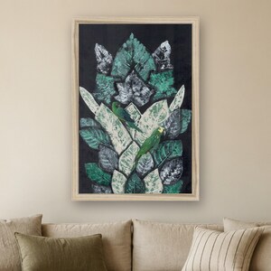 May include: A framed rectangular wall art piece featuring a dark black background with a central vertical arrangement of layered green and white leaves. Two small green parrots with yellow accents are perched among the foliage.