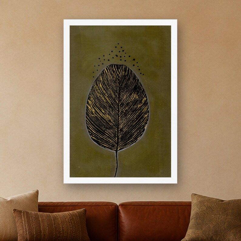 May include: A rectangular wall art print featuring a large, stylized black leaf with visible gold-toned vein details centered on a muted olive green background. Above the leaf, a small arc of black dots is arranged in a triangular pattern.