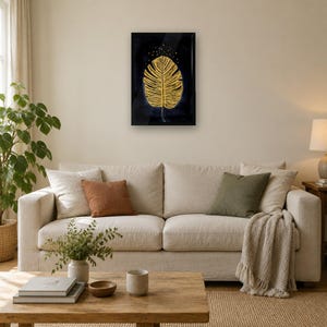 May include: A rectangular wall art piece featuring a large, textured gold leaf illustration centered against a deep black background, accented with small gold dots above the leaf, all enclosed within a thin black frame.