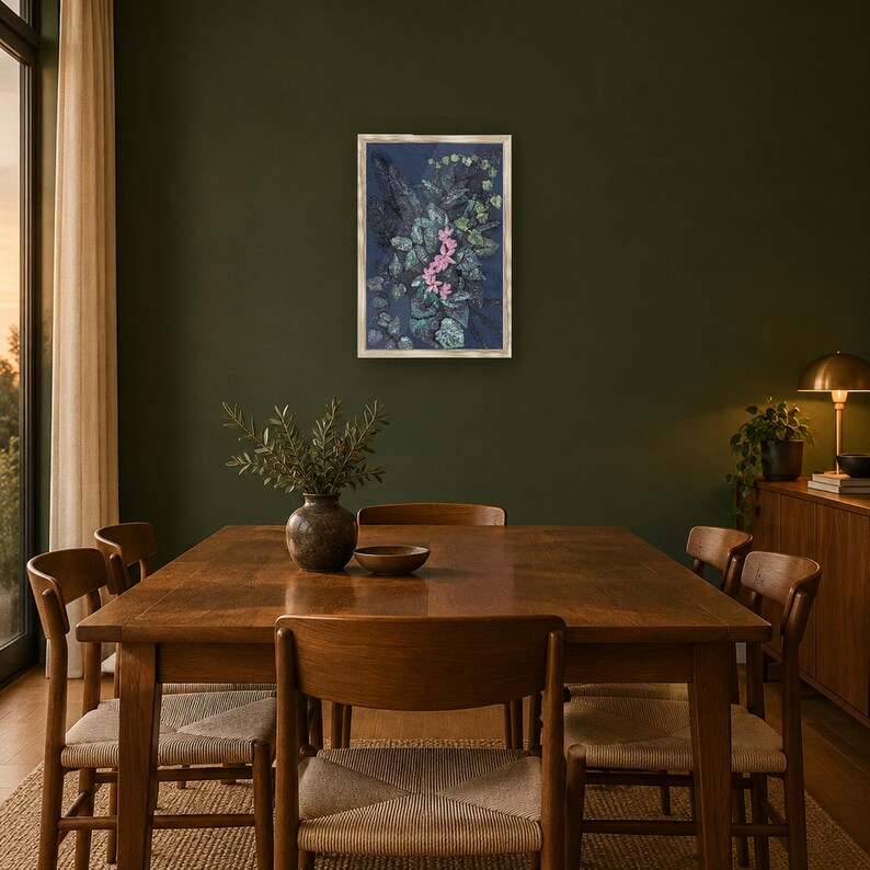 May include: A rectangular framed wall art piece featuring a dark blue background with a dense arrangement of green foliage and a cluster of small pink flowers in the center, enclosed in a light-colored wooden frame hanging on a dark wall.