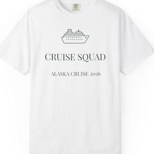 Può includere: Una t-shirt bianca a maniche corte con un disegno a linee nere di una nave da crociera centrato in alto. Sotto la nave, il testo recita CRUISE SQUAD in grassetto, seguito dal testo ALASKA CRUISE 2026 in un carattere più piccolo e sottile.