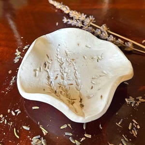 Puede incluir: Un cuenco de cerámica blanca de forma irregular con un patrón de rama de lavanda en relieve en su superficie interior. El plato tiene un acabado mate liso con bordes suavemente ondulados y un centro cóncavo poco profundo.