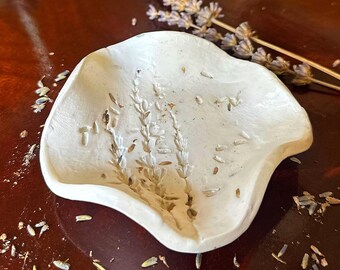 Könnte beinhalten: Eine weiße, unregelmäßig geformte Keramikschale mit einem geprägten Lavendelzweig-Muster auf der Innenseite. Die Schale hat eine glatte, matte Oberfläche mit sanft gewellten Rändern und eine flache, konkave Vertiefung.