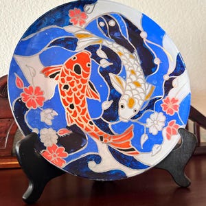 Puede incluir: Plato decorativo con diseño de carpas koi. El plato tiene un fondo azul con olas blancas y carpas koi rojas y blancas. También hay detalles florales rosas y blancos. El plato se exhibe sobre un soporte negro.