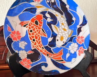 May include: Decorative plate with a koi carp design. The plate has a blue background with white waves and red and white koi carp. Pink and white floral accents are also present. The plate is displayed on a black stand.