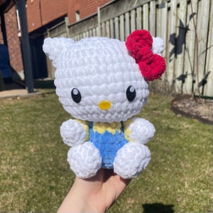 Peut inclure: Peluche Hello Kitty en crochet blanc avec un nœud rouge et une salopette bleue.