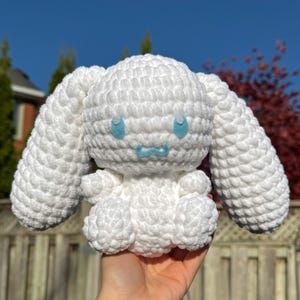 May include: A white crochet plush toy with large, floppy ears and a rounded body. The toy has light blue eyes and a small, light blue mouth. The plush is held in a hand, showing its size and texture. The toy is handmade.
