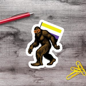 Pode incluir: Um autocolante recortado com uma criatura Bigfoot castanha e peluda a usar óculos de sol cor de laranja em forma de estrela. A criatura segura uma bandeira com riscas horizontais amarelas, brancas e roxas. O texto APHANTASTICDESIGNS está impresso na perna.