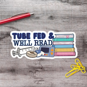 Op de afbeelding: Een witte sticker met de tekst "TUBE FED & WELL READ" in blauw. De sticker bevat illustraties van een spuit, medische apparaten en een stapel kleurrijke boeken. Een rode pen en gele paperclips zijn ook zichtbaar.