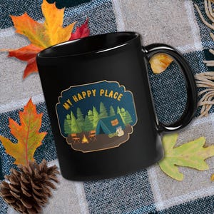 Könnte beinhalten: Ein schwarzer Keramikbecher mit einer grafischen Illustration einer Campingszene mit Zelt, Lagerfeuer und Tannenbäumen unter einem Sternenhimmel. Der Text "MY HAPPY PLACE" ist in gelben Buchstaben über der Campingszene aufgedruckt.